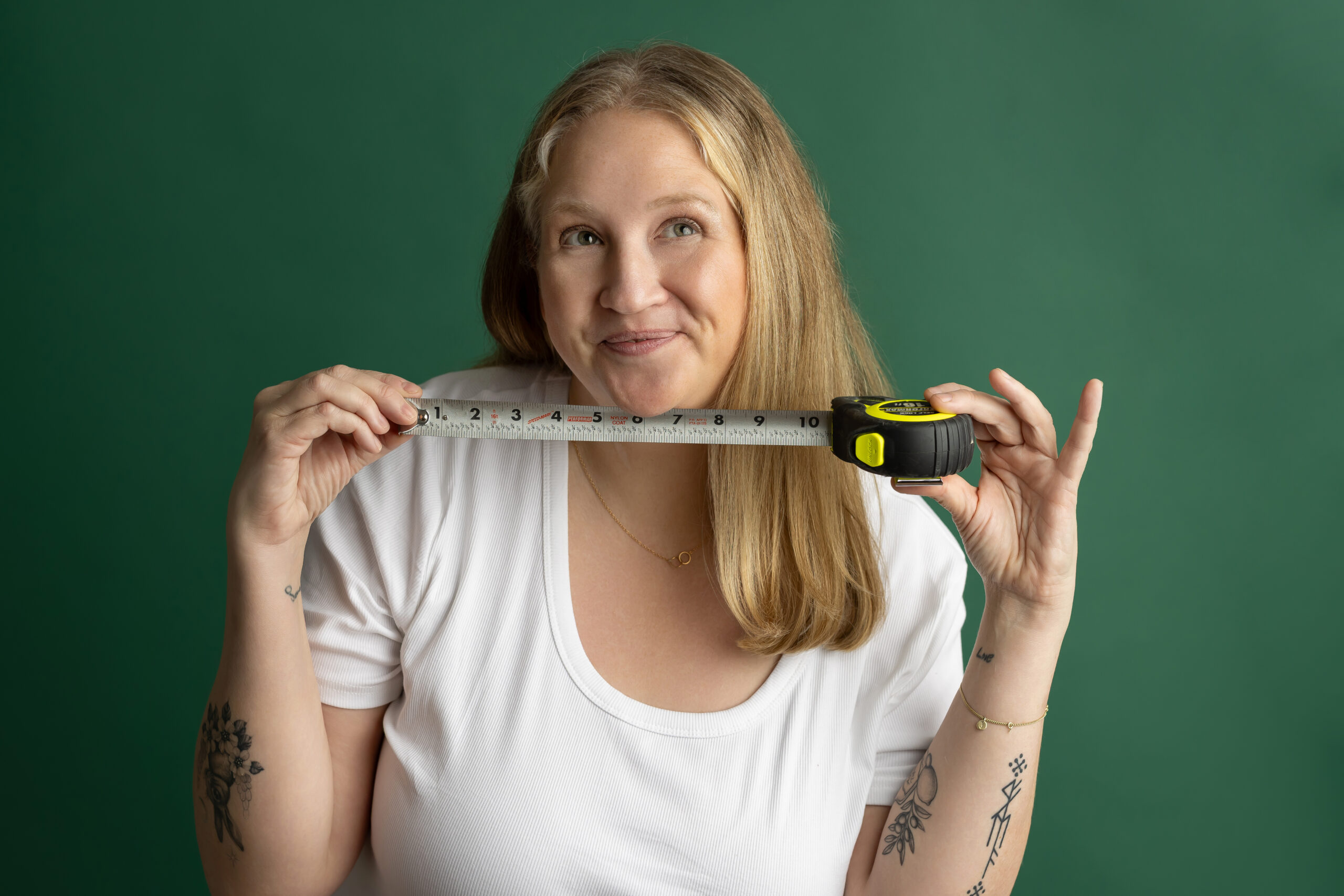 Smiling woman in a white shirt holds a measuring tape horizontally across her chest against a green background, as if measuring.