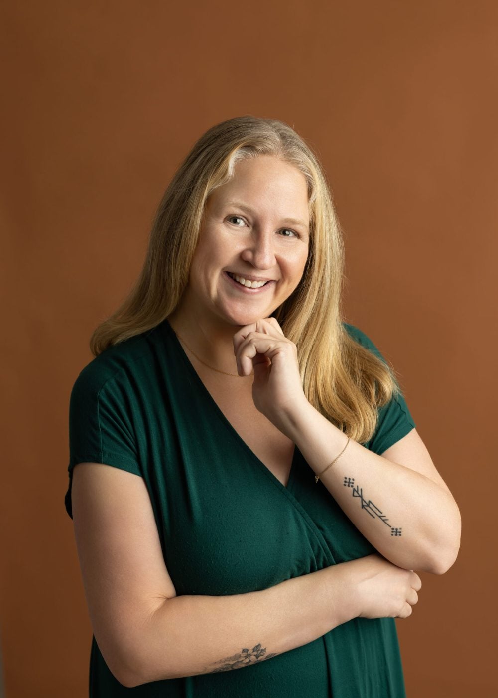 Smiling woman with long blonde hair in a dark green V-neck dress, posed against a rust-colored background