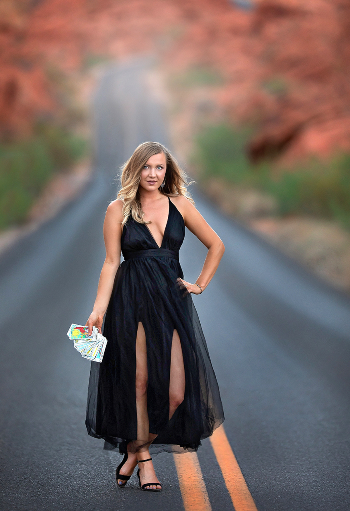 Woman in a black dress stands on a road with orange lane lines, holding a fan of cards in one hand.